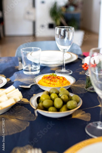 Obraz Dining table with water glasses, a bowl of hummus, and olives, decorated with cotton branches on a blue and gold cloth. Christmas table setup with olive and mediterranean food.