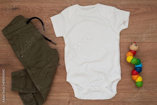 Fototapeta Mockup of white baby bodysuit on wood background with pants and colorful toy. Blank baby clothes template mock up, flat lay styled stock photo.