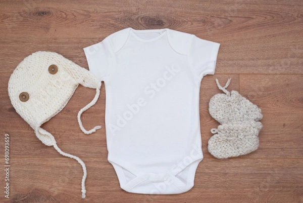 Fototapeta Mockup of white baby bodysuit on wood background with knitted booties and hat. Blank baby clothes template mock up.
