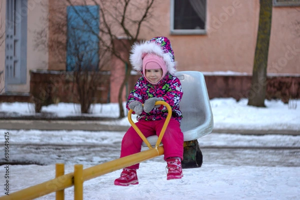 Obraz A little girl in a warm pink winter jacket rides on a swing at the playground