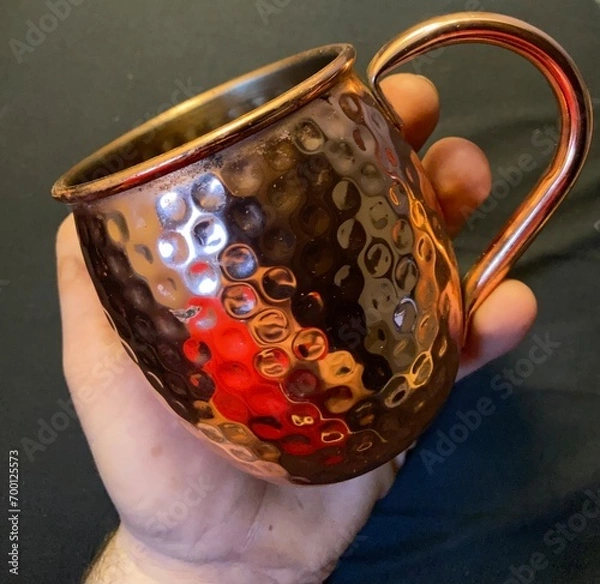 Obraz mug, cup in red galvanized copper metal with handle. Hand holding glass for Moscow Mule Cocktale. Image with neutral and light background.