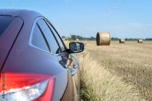 Fototapeta Close up view red maroon crossover car on freshly harvested field with straw bales. Selective focus.