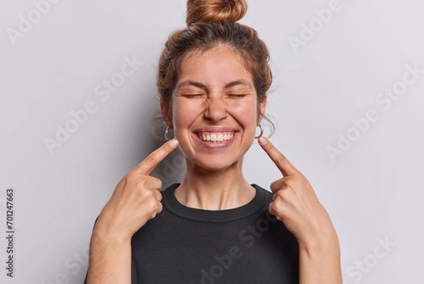 Fototapeta Photo of pleasant looking cheerful woman points index fingers at toothy smile shows wwell cared whie even teeth dressed in casual black t shirt isolated over whtie studio background feels happy