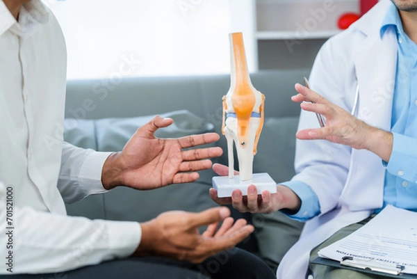 Fototapeta male patient and male doctor discussing a model of a knee joint, likely focusing on condition of knee arthritis during a medical consultation.