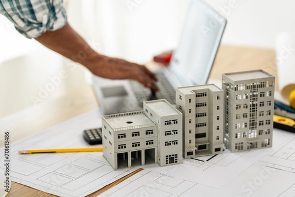 Fototapeta Close up Asian individual architect or engineer holding building scale model of multi-story building. design or review of architectural plans laid out on work surface in front of them.