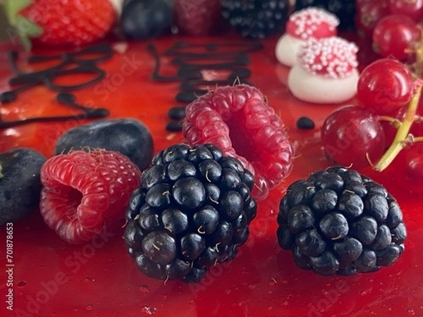 Fototapeta Cake decoration close up, blackberries in the forefront and other beautiful berries and sugar decoration on a vibrant red glaze. 