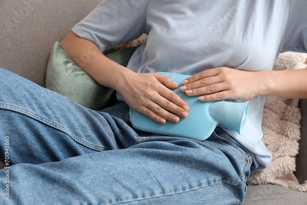 Obraz Young woman with hot water bottle sitting on sofa and suffering from abdominal pain at home
