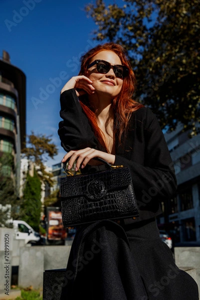 Obraz Young and beautiful model poses for the camera. Red-haired model posing for the camera with her crocodile leather, black, luxury bag outside. Fashion photography