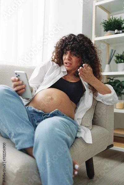 Fototapeta Pregnant woman blogger sits on the couch at home and takes pictures of herself on the phone, selfie and video call, consultation with the doctor online, pregnancy management, freelance work