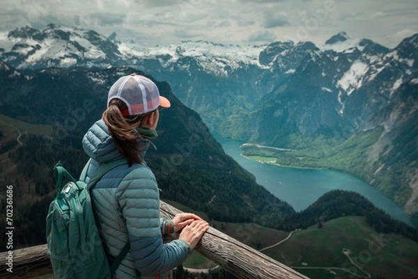 Fototapeta Female hiker with backpack looking at turquoise lake Königssee from above viewpoint of mountain peak Jenner at Berchtesgaden Bavaria, snow-capped mountains in the background.