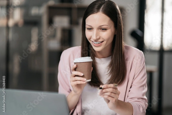 Fototapeta businesswoman sits in a office working on laptop and enjoys a coffee.