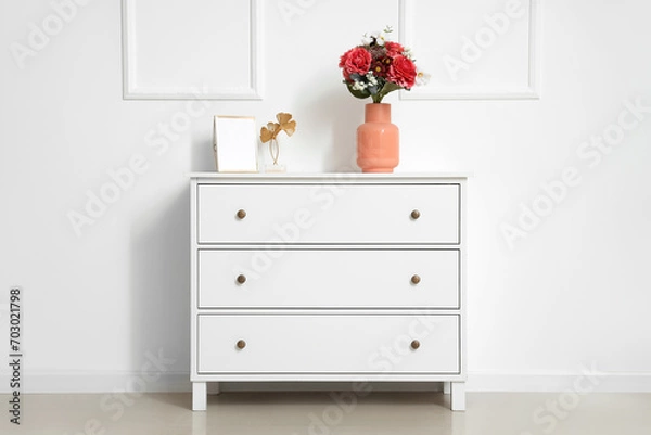 Fototapeta Vase with beautiful artificial flowers on chest of drawers near white wall