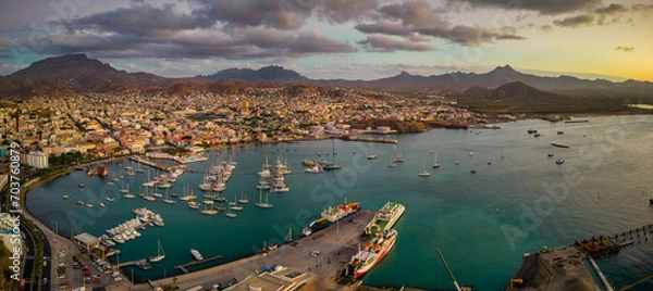 Obraz Panoramic aerial view of Mindelo city at sunset, with the marina and boats in the foreground, surrounded by the vibrant cityscape and mountains in the background under a warm, glowing sky