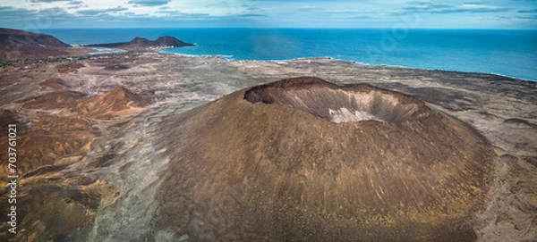 Obraz Aerial view of a coastal volcanic landscape (Viana volcano, Sao Vicente) with a distinct crater in the foreground, sparse vegetation, and a small village by the sea, under a cloudy sky