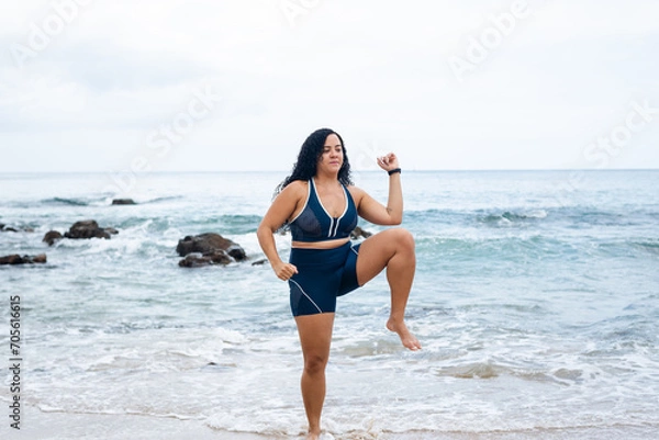 Fototapeta Fitness woman, standing, doing leg exercises facing the camera on the beach sand against the sea and cloudy sky.