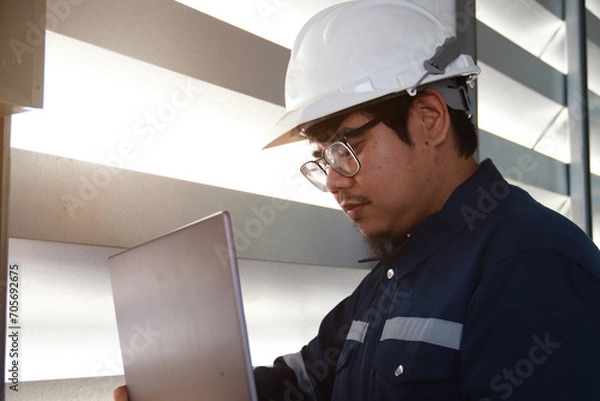 Obraz Chief engineer in hard hat walks through modern factory lighting while holding laptop. Successful handsome man in modern industrial environment.