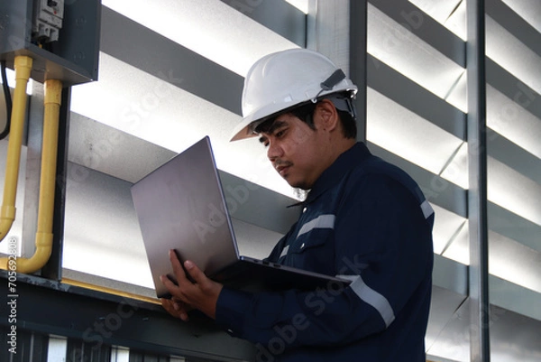 Obraz Chief engineer in hard hat walks through modern factory lighting while holding laptop. Successful handsome man in modern industrial environment.
