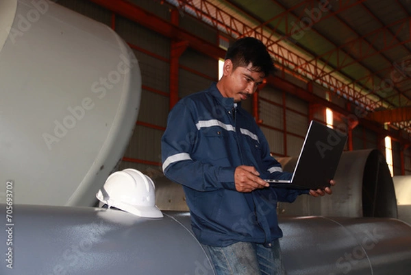 Obraz Chief engineer in hard hat walks through modern factory lighting while holding laptop. Successful handsome man in modern industrial environment.