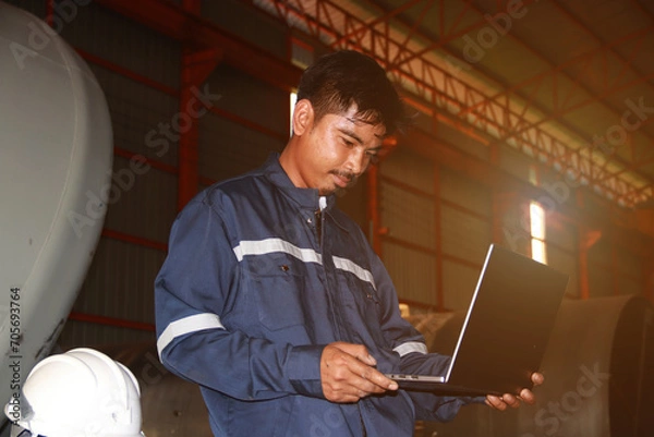 Obraz Chief engineer in hard hat walks through modern factory lighting while holding laptop. Successful handsome man in modern industrial environment.