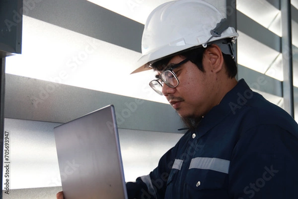 Obraz Chief engineer in hard hat walks through modern factory lighting while holding laptop. Successful handsome man in modern industrial environment.