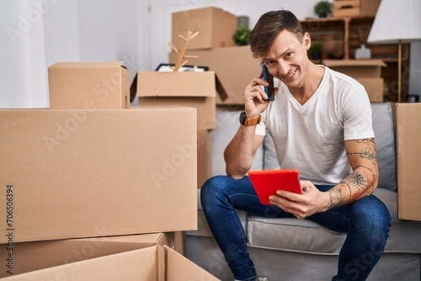 Fototapeta Young man talking on smartphone using credit card at new home