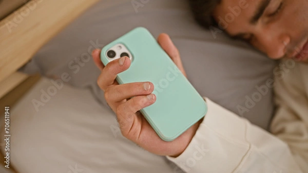 Fototapeta Exhausted young hispanic man, holding his phone, deeply asleep, resting comfortably in a cozy bed at home, with his whole room as a relaxed background.