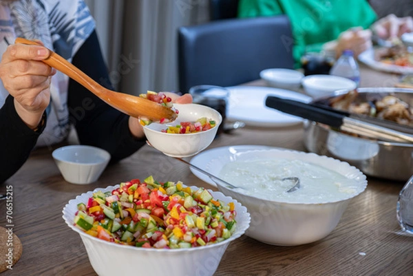 Fototapeta The wife fills a salad bowl with a large bowl of yogurt next to it to provide the guests with what they need at lunchtime at the large collective table for the large family.