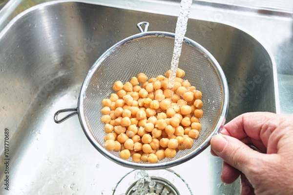 Fototapeta Pre-cooked canned chickpeas are rinsed with water in a sieve over the sink before being used in various dishes, healthy legumes are rich in protein, fiber and minerals, copy space