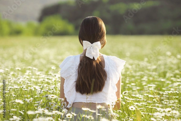 Fototapeta Ponytail faceless woman hairstyle with white bow clip on long brown hair. A young girl from the back sits in a field of daisies and enjoys freedom and calm. Mental health. Chamomile field landscape.