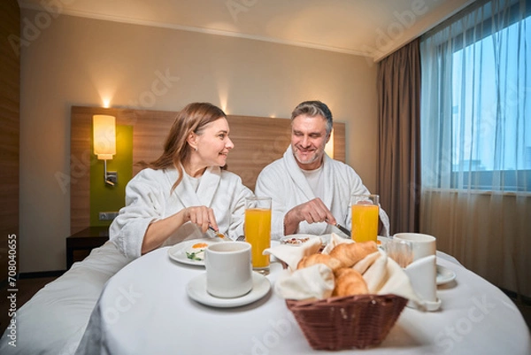 Fototapeta Smiling adult caucasian couple having breakfast on bed at table in hotel room