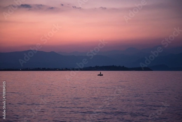 Obraz A lonely fisher standing on an inflatable boat with an oar against a backdrop of misty mountains and a Garda lake