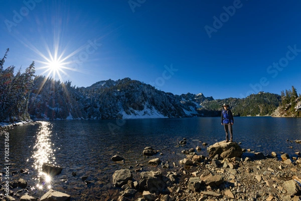 Fototapeta Adventurous athletic male hiker standing on a hill overlooking an alpine lake and tall mountains, on a beautiful sunny day in the Pacific Northwest.
