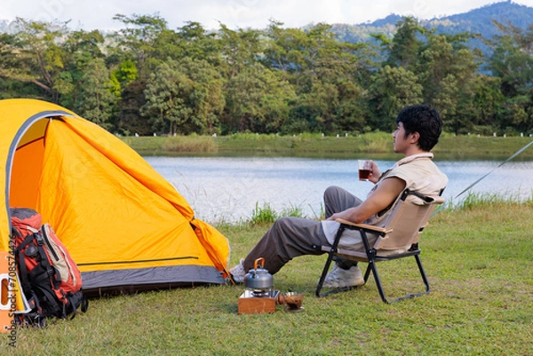 Fototapeta An Asian man sitting and drinking coffee near camping tent beside the lake and mountain in the sunshine morning with backpack and mobile solar cell