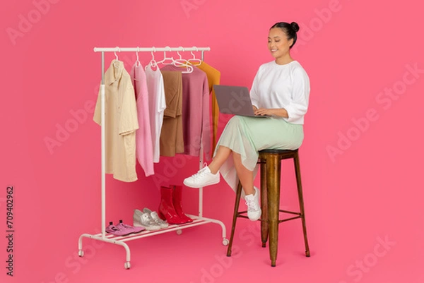 Fototapeta Beautiful asian woman sitting with laptop near clothing rack, managing online boutique