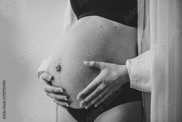 Fototapeta Closeup belly of a woman. Pregnancy motherhood procreation concept. Pregnant female waiting for newborn baby. Young girl touching and holding belly, caring about health indoors. Black and white photo