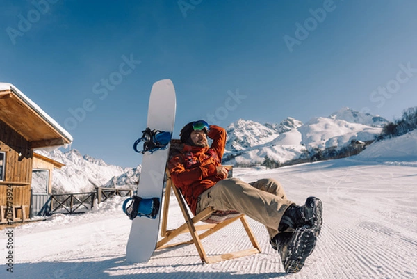 Fototapeta snowboarder sitting in sun lounger on sunny winter day, corduroy of prepared downhill slope at ski resort