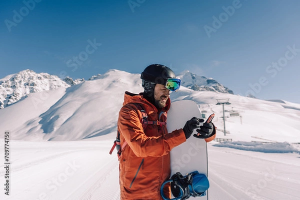 Fototapeta snowboarder holding snowboard and smartphone in his hands on prepared ski slope communicates by phone online and answers messages