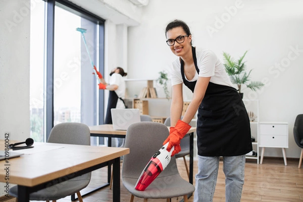 Fototapeta Happy smiling multicultural employees of company cleaning in bright, spacious modern office. Multiethnic young female cleaner vacuuming chair with portable cordless vacuum cleaner, looking at camera.