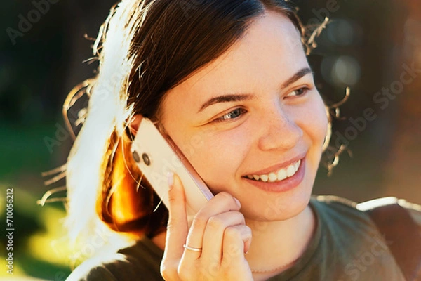 Obraz Happy, smiling young girl talking on a mobile phone in a city park. Young beautiful lady talking on her smartphone outdoors. An attractive girl happily communicates with a friend. Communication