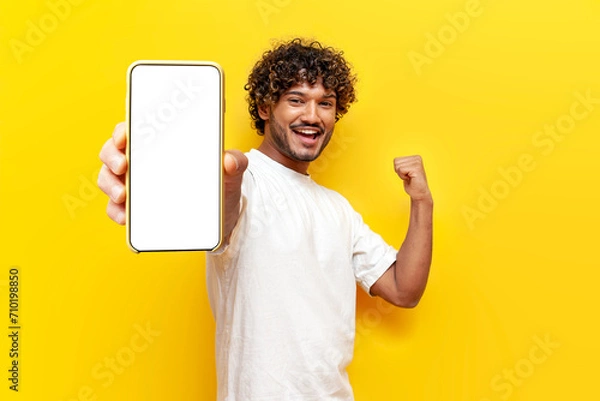 Fototapeta young curly indian man shows blank smartphone screen and rejoices in victory on yellow isolated background, guy wins online and celebrates success with phone