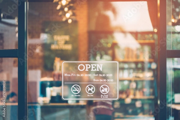 Fototapeta Stickers showing opening and closing times. Close up an open sign on the front window of a coffee shop/cafe and a blurred, bokeh image inside the shop.