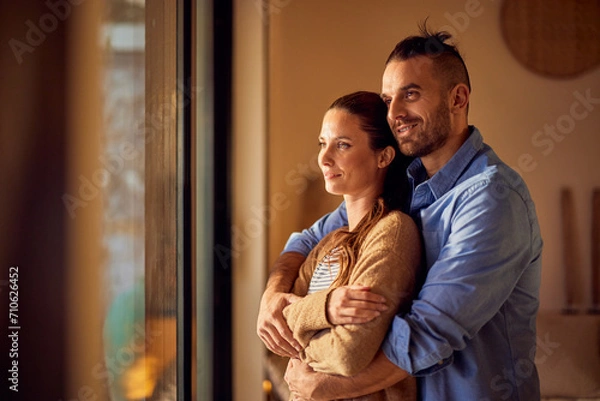 Fototapeta Two lovers in a hug, standing and looking through the window, smiling.