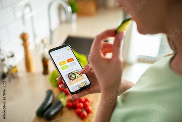 Fototapeta Woman using a meal planning app on smartphone in kitchen