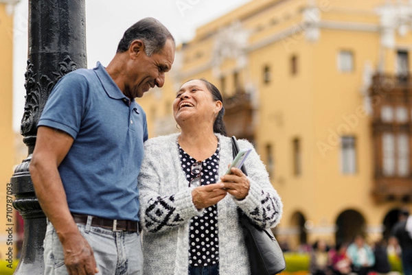Fototapeta Multiracial adult couple happily using their mobile phones while strolling through the city on a beautiful day. Copyspace