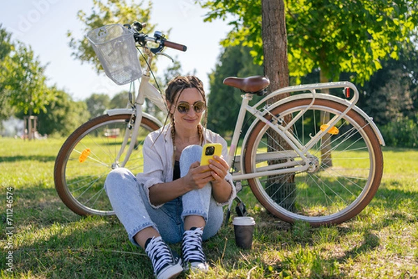 Fototapeta Smiling young hipster woman with bicycle using mobile phone in city park. Talking by video call, using internet