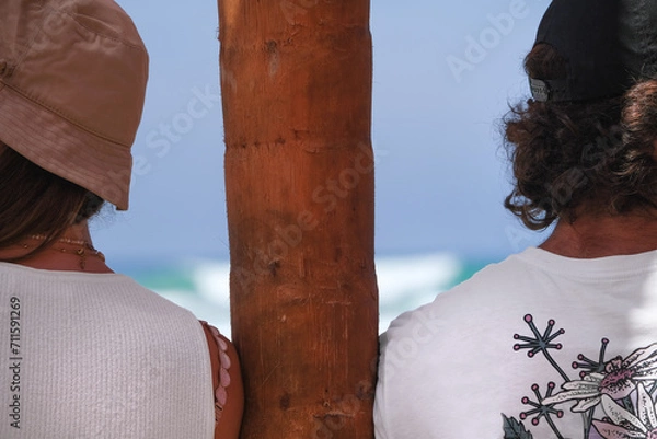 Fototapeta Man and a woman lean against a palm tree and look in different directions against the backdrop of the ocean, rear view