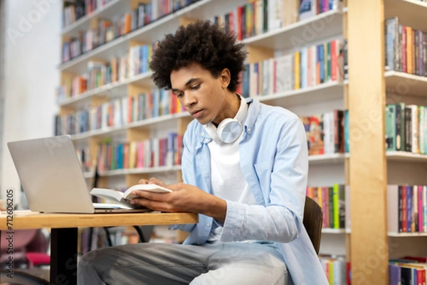 Obraz Focused black student guy sitting in library in front of laptop, diligently reading book, creating scene of concentrated study and blended learning methods