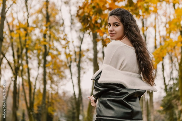 Fototapeta Attractive pretty woman walking outdoor, portrait of young lady in warm sunny autumn park season, fall, yellow orange red leaves, dressed leather wool fur jacket smiling having fun 
