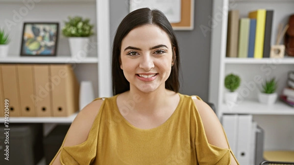Obraz Smiling young woman in casual office attire seated indoors with a neat shelf background.