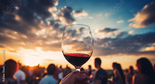 Fototapeta Hand holding a glass of red wine, capturing the essence of a relaxed social evening at sunset.